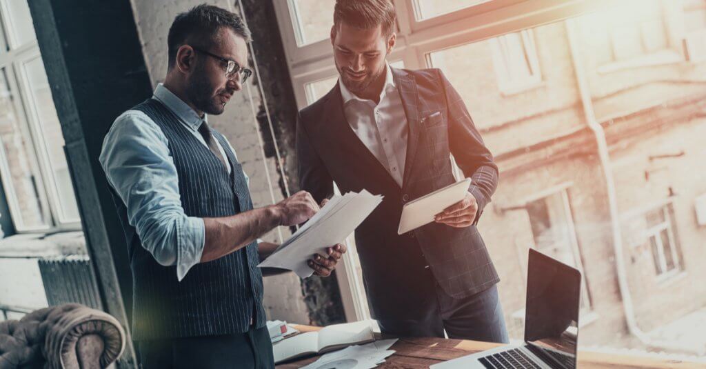 Two men in suits stand beside a laptop, discussing details related to Capital Premium Financing.