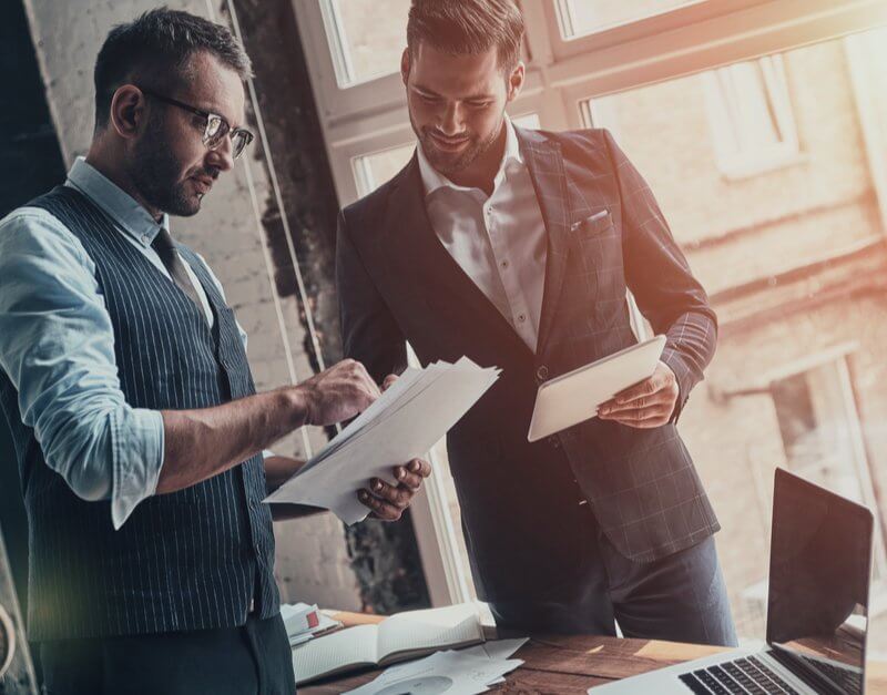 Two men in suits stand beside a laptop, discussing details related to Capital Premium Financing.