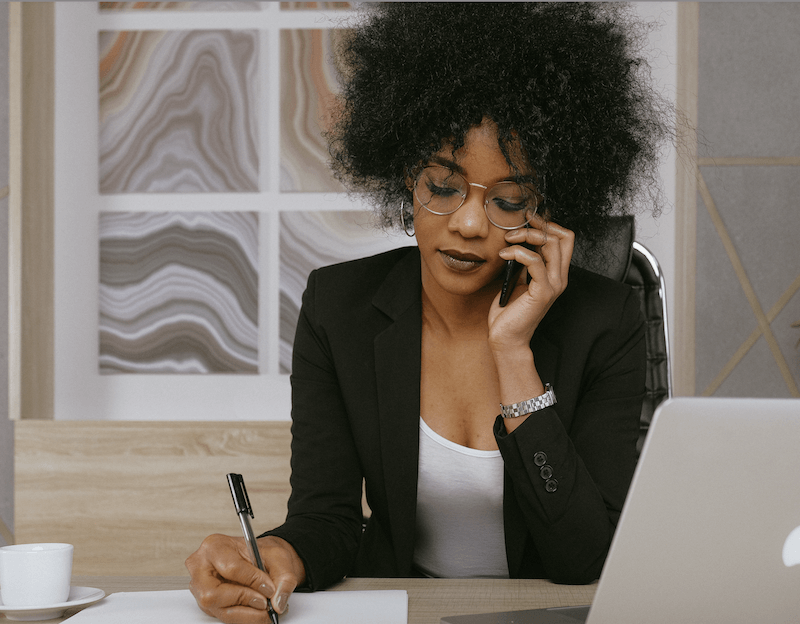 A woman in a business suit writes on paper while talking on the phone, representing a professional work environment.