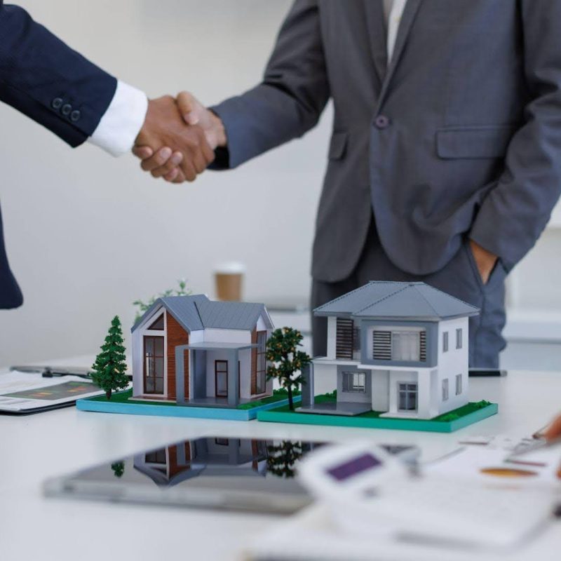 Two businessmen engage in a handshake above a house model, representing a real estate partnership