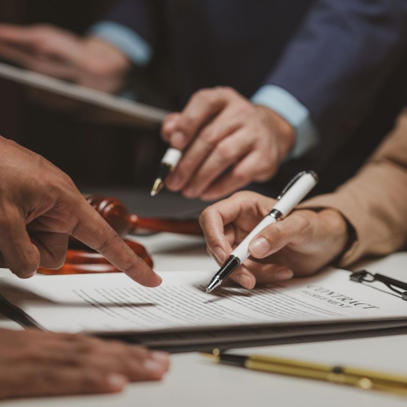 Close-up of hands during a contract signing one person pointing to a specific section on the agreement document while another prepares to sign with a pen, with a gavel visible in the background suggesting a legal or formal context.
