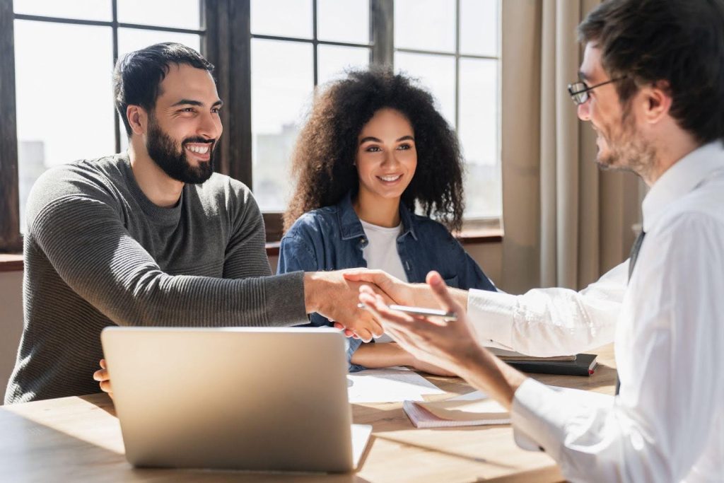 Three people shaking hands at a table with a laptop, suggesting collaboration