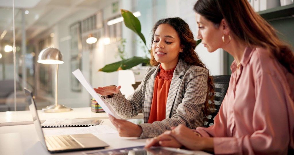 Two women work together on paperwork at a desk in an office