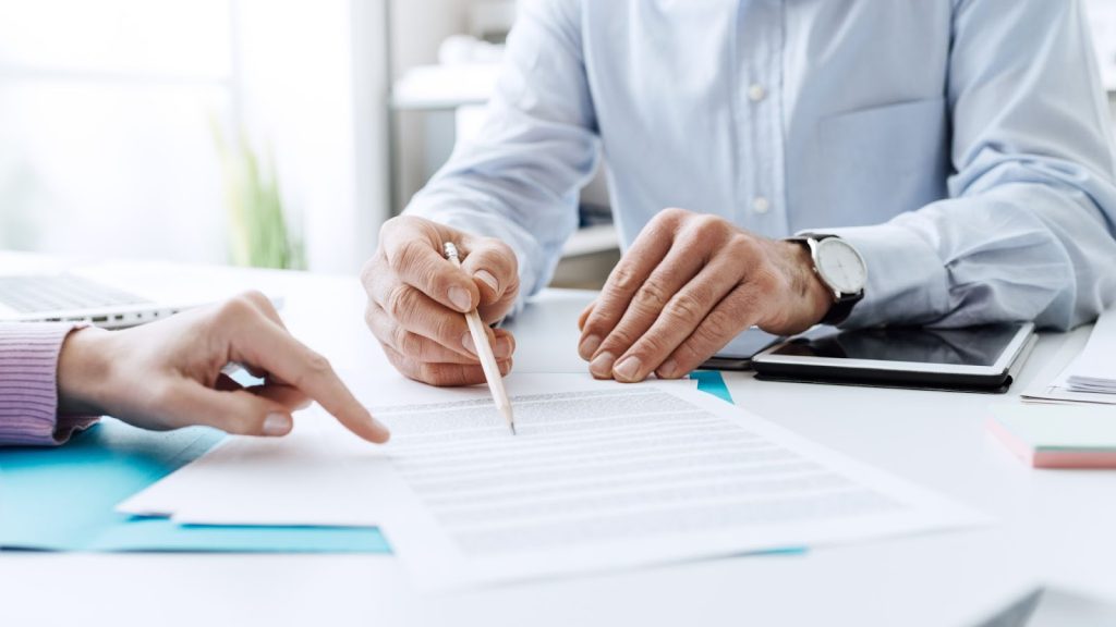 Two people reviewing documents at a desk with a laptop and tablet.
