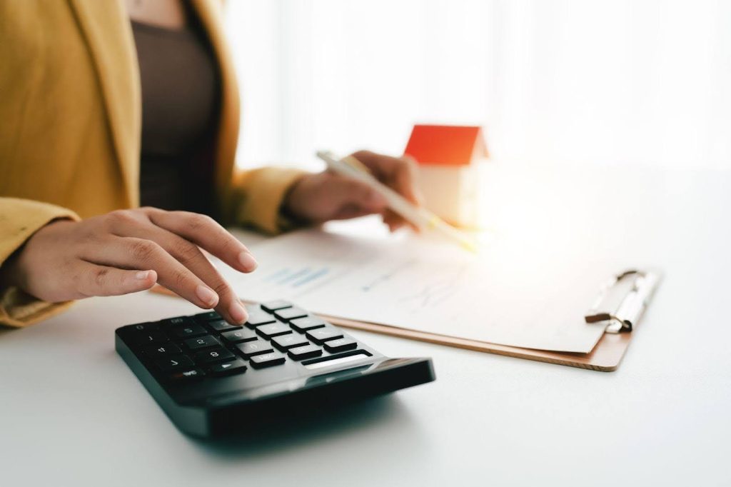 A woman intently using a calculator to determine the value of a house surrounded by paperwork and a laptop