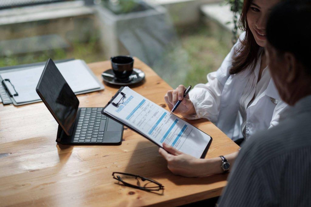 A woman sits at a table with a laptop while a man observes her attentively