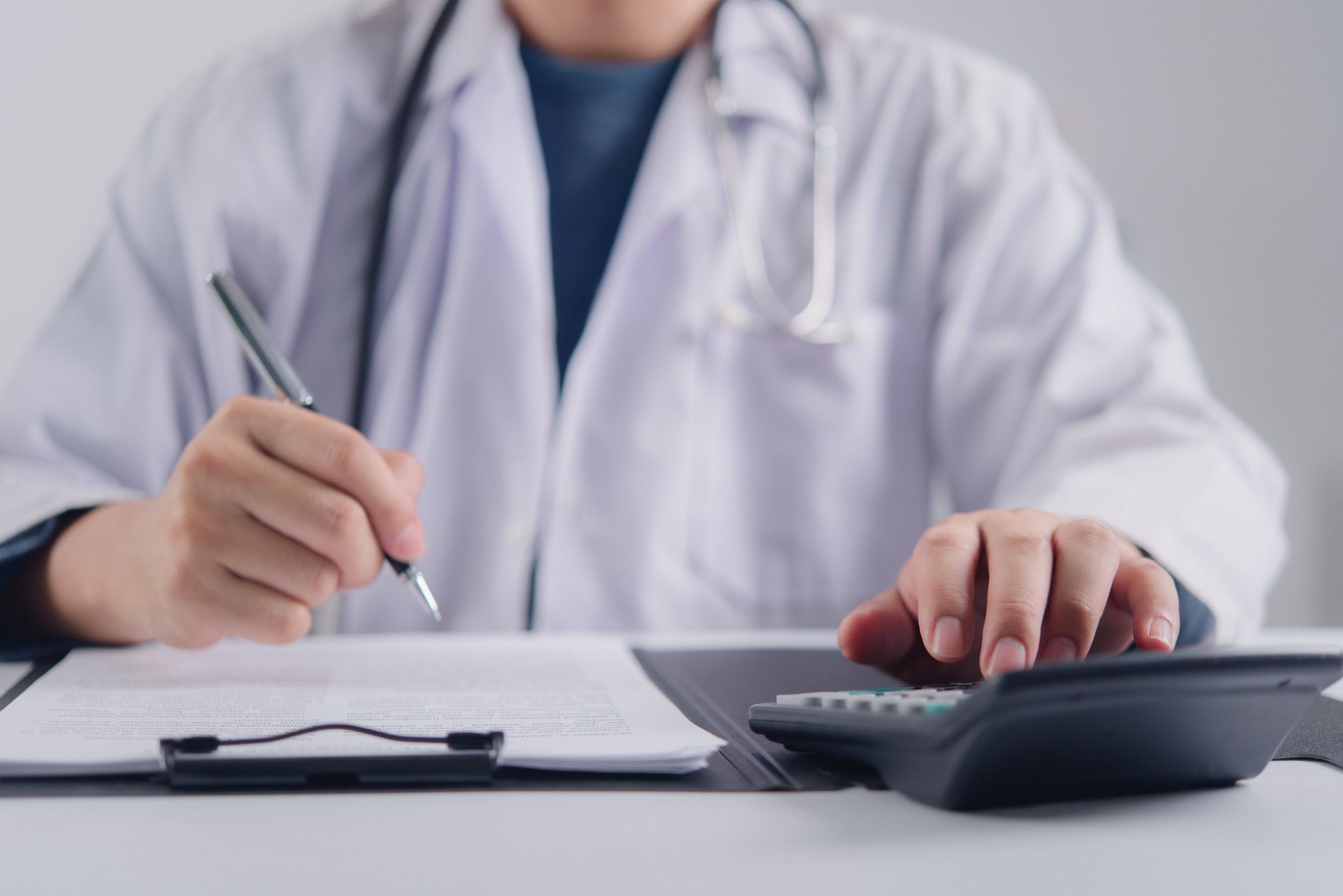 A close-up of a doctor writing on a clipboard while using a calculator.