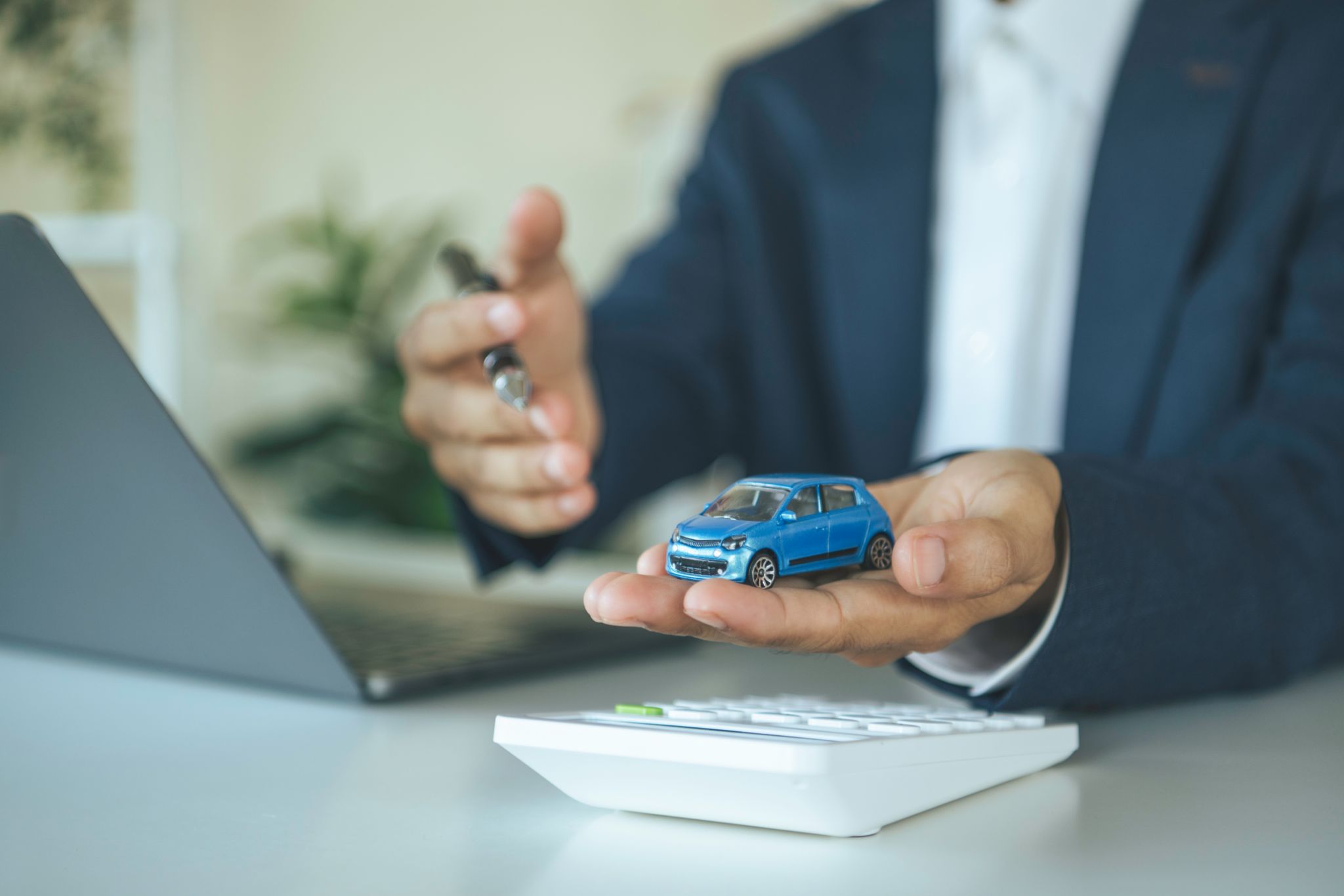A person in a suit holding a small blue toy car over a calculator at a desk.