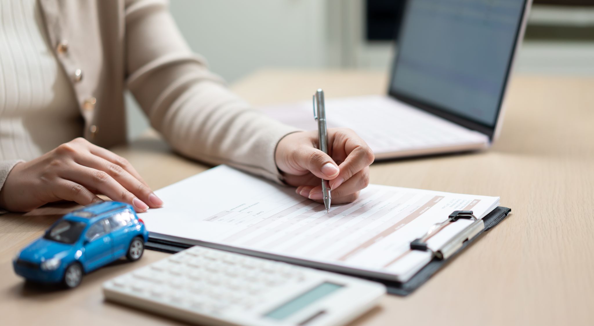 Person filling out car loan or insurance paperwork with a pen beside a toy car and calculator.